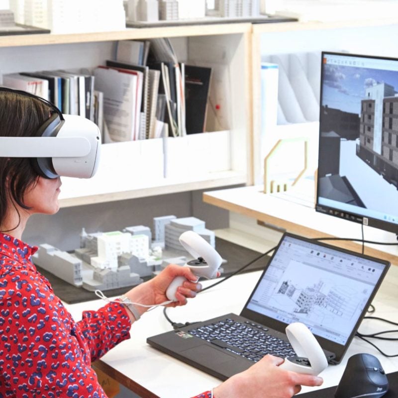 MDLR Architects — A woman wearing VR goggles and holding controllers stands in the MDLR office. She interacts with architectural models displayed on two screens, explor